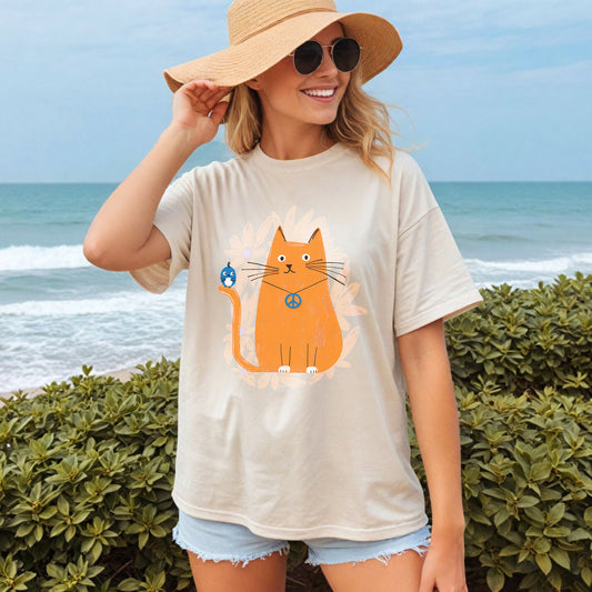 Woman wearing an ivory t-shirt featuring on the front an orange cat wearing a "peace" sign necklace; perched on the cat's tail is a cute blue bird. A spray of pale orange flowers in the background.